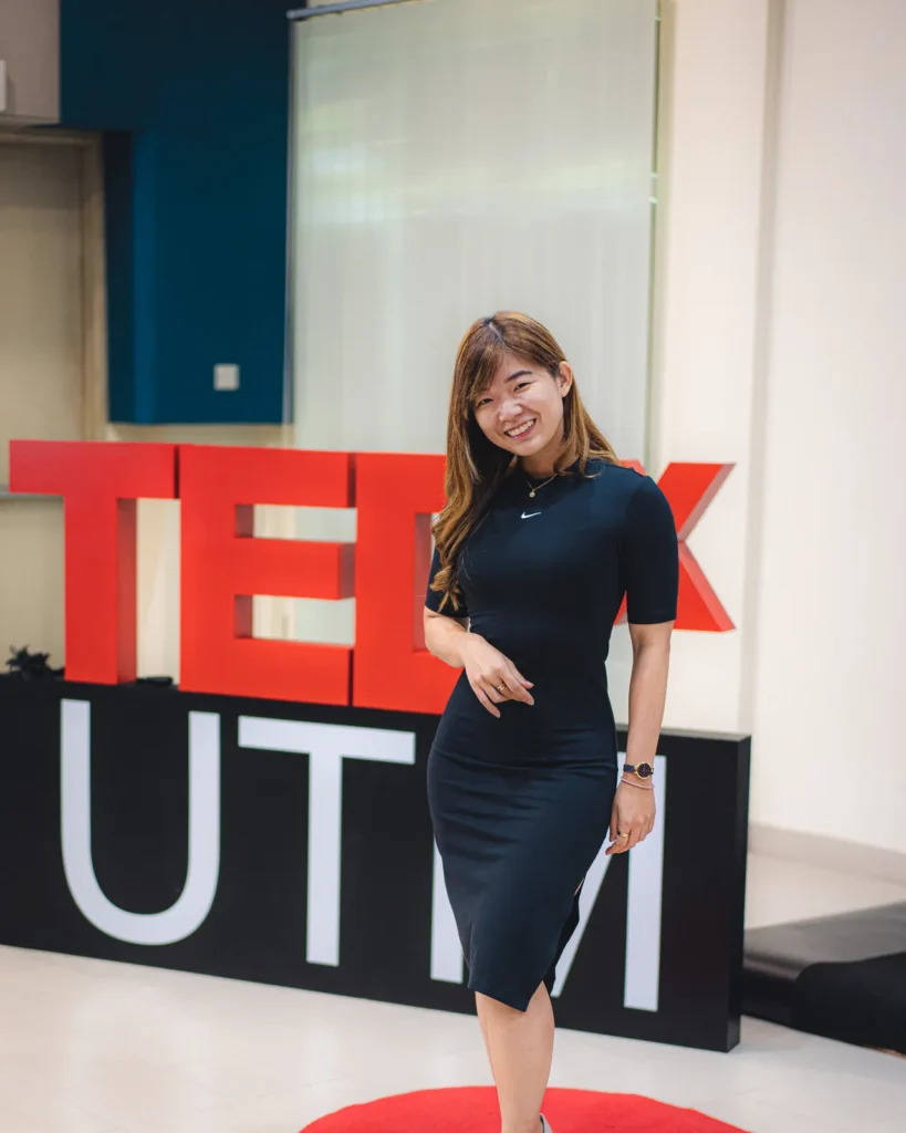 Yin Sinn standing on stage at TEDxUTM, smiling in a navy dress